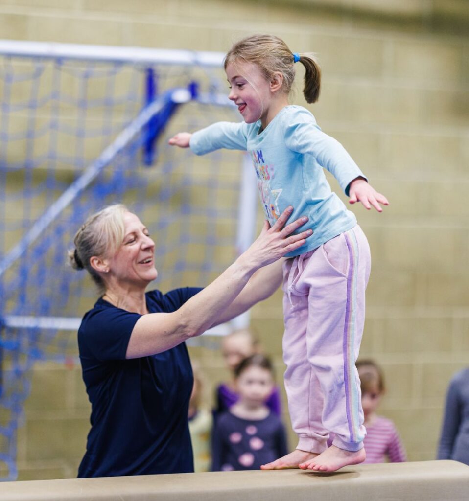 young girl on gymnastics beam with coach supporting her